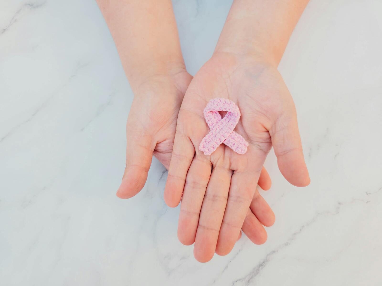 Pink ribbon in hands symbolizing breast cancer awareness on marble background.
